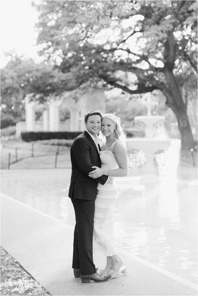 black and white photo with a couple in front of a fountain smilng at the camera